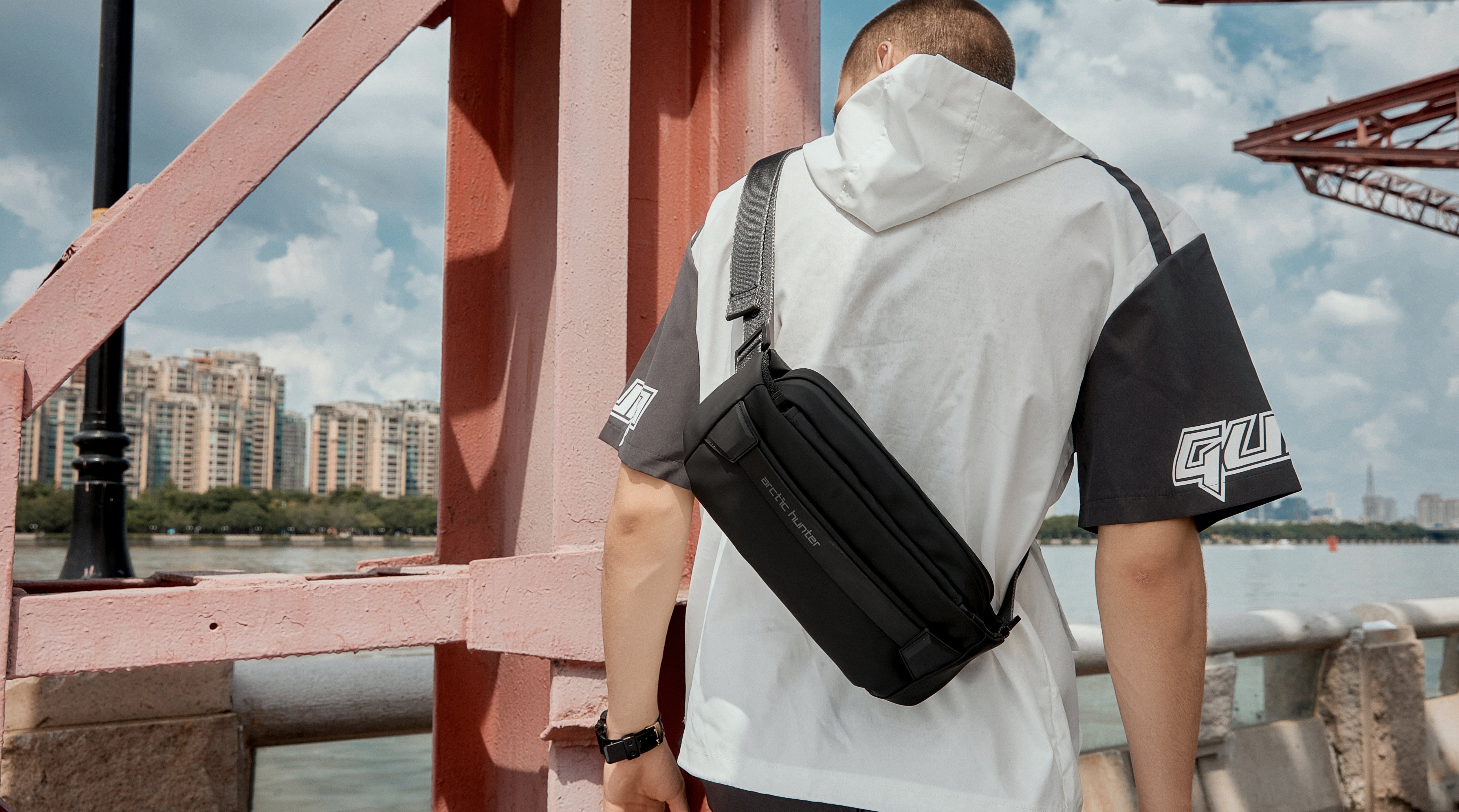 Moderner Mann trägt die schwarze Umhängetasche - Arctic Prox Crossbody im urbanen Outdoor-Look auf einer Brücke – ideal für Stadtentdecker.