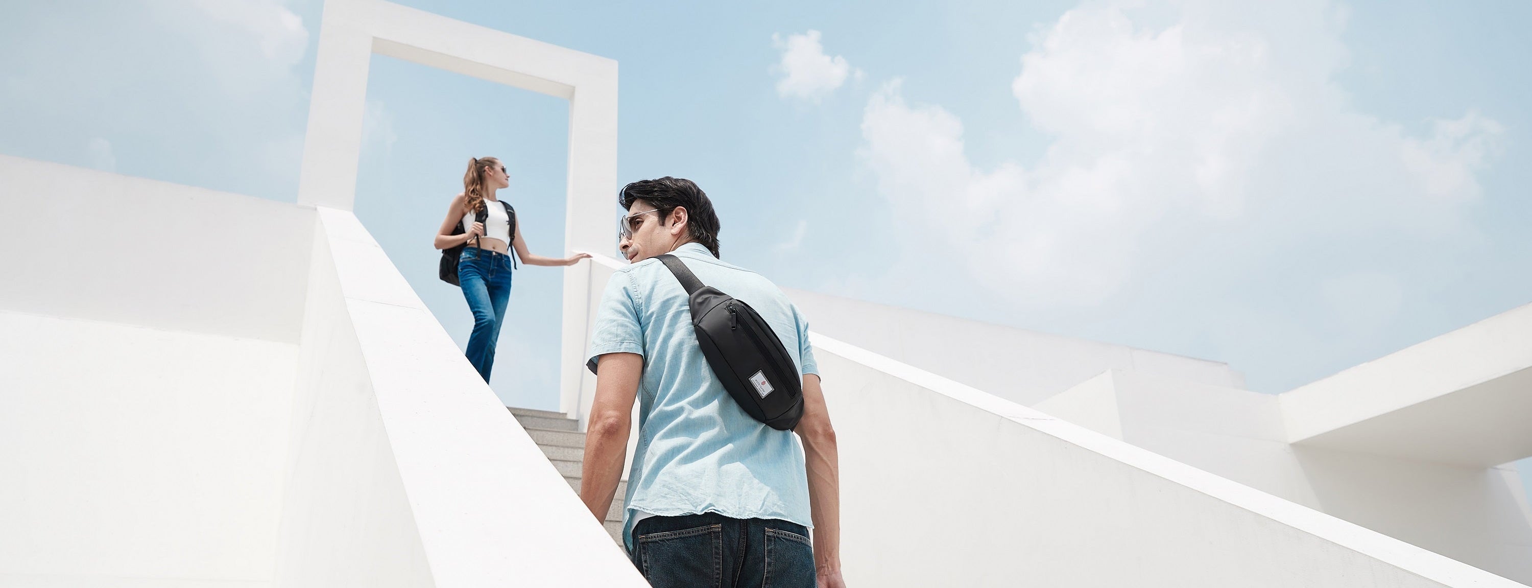 Ein Mann trägt eine Crossbody Tasche auf einer Treppe, während eine Frau vor ihm die Treppe hinaufgeht. Beide sind im urbanen Alltag unterwegs.