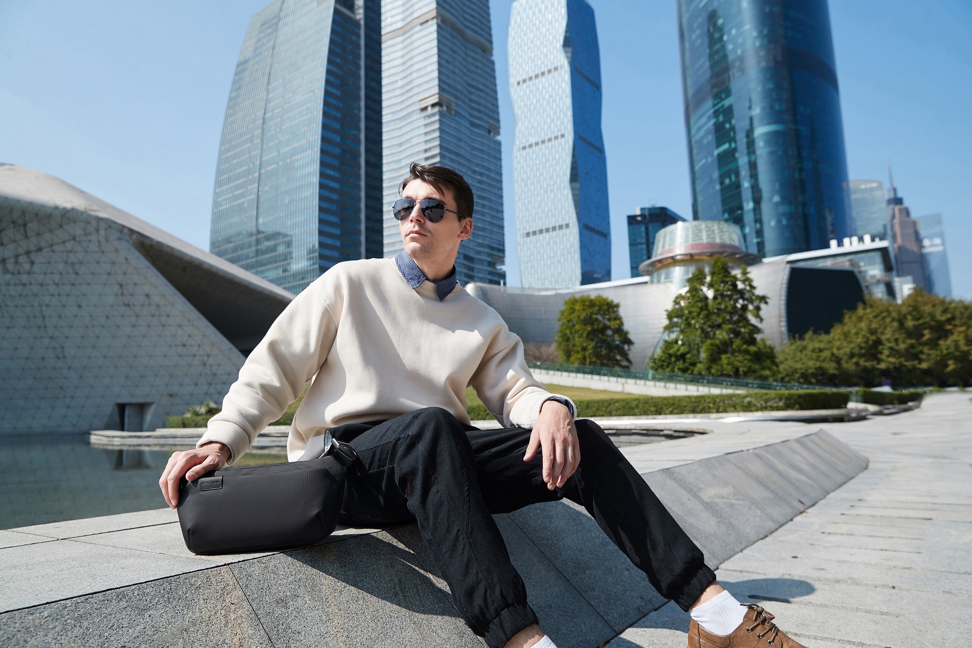 Moderner Mann mit schwarzer Umhängetasche sitzt vor moderner Skyline in der Stadt – stylischer urbaner Look für Alltag, Reisen und Business.

