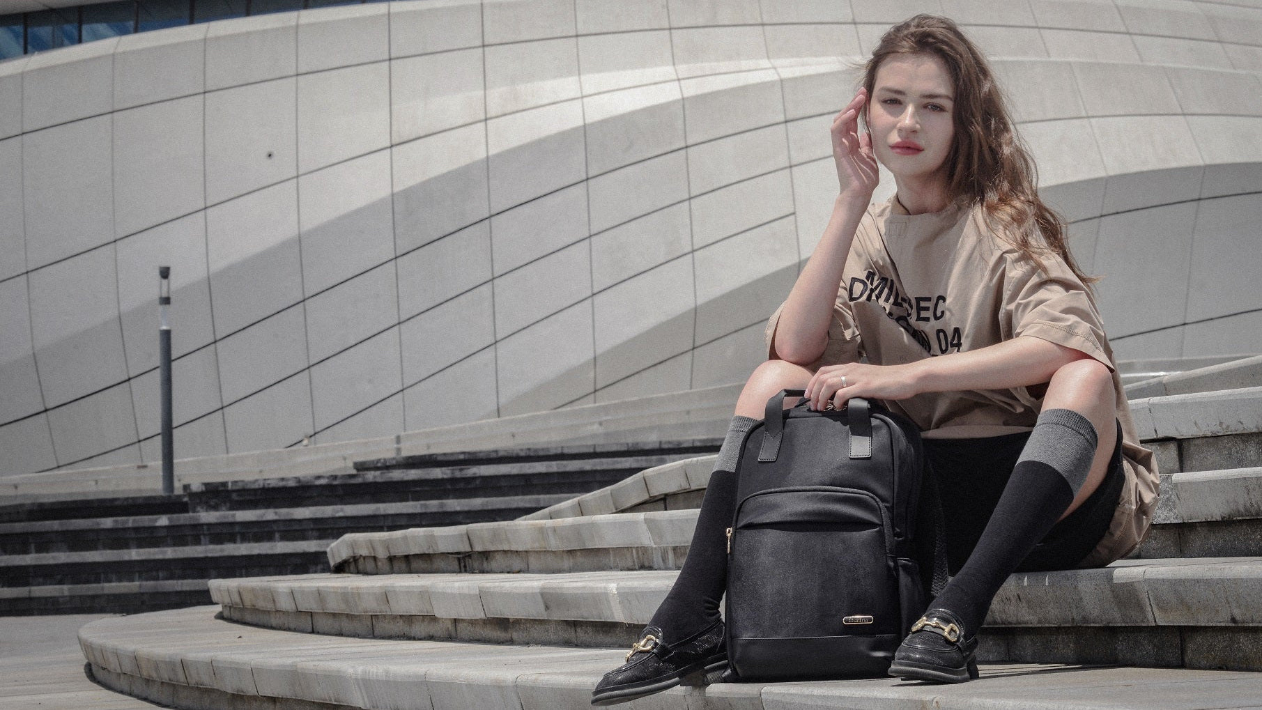 Frau sitzt auf Treppe in der Stadt und trägt urbanen Damen-Rucksack, modernes Gebäude im Hintergrund